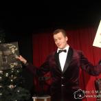 Ein Mann in Samtanzug steht auf einer Bühne neben einem Weihnachtsbaum und hält verschiedene Bilder.