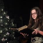 Eine junge Frau liest in einem alten Buch, während im Hintergrund ein beleuchteter Weihnachtsbaum zu sehen ist.
